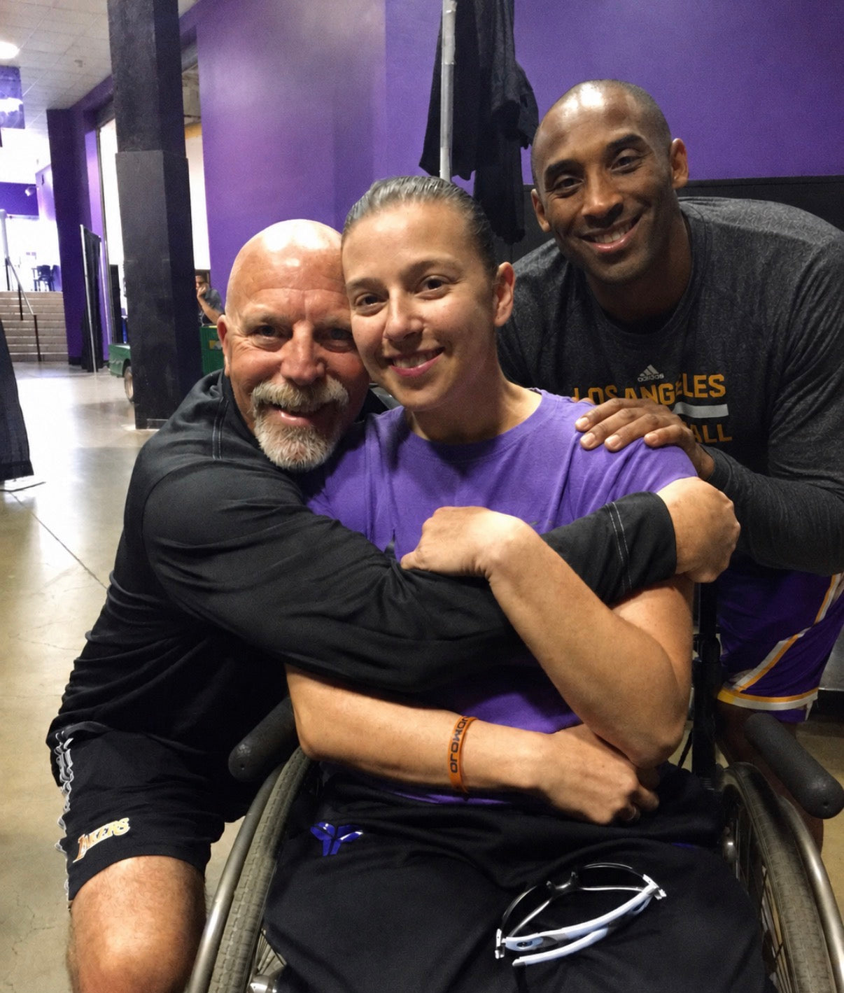 A group photo in an arena hallway featuring Joan Rubinger in her wheelchair wearing a purple shirt, surrounded by Lakers' trainer Gary Vitti on the left, and Kobe Bryant on the right.