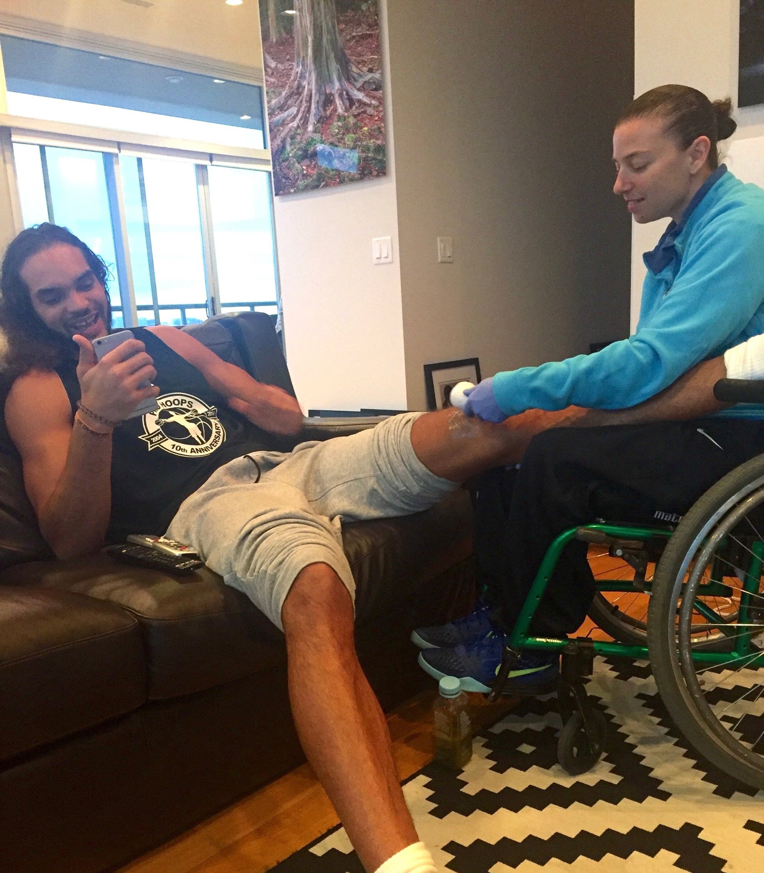 Former NBA player Joakim Noah in a black tank top and light gray shorts reclines on a brown leather couch while Joan Rubinger, in a bright blue top, attends to his leg, which is wrapped in white bandages. The man is smiling and giving a thumbs-up.