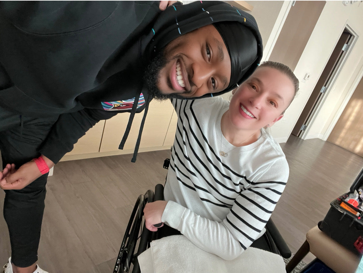 NFL Linebacker Owen Pappoe, standing and leaning over Joan Rubinger in her wheelchair, smiling indoors.